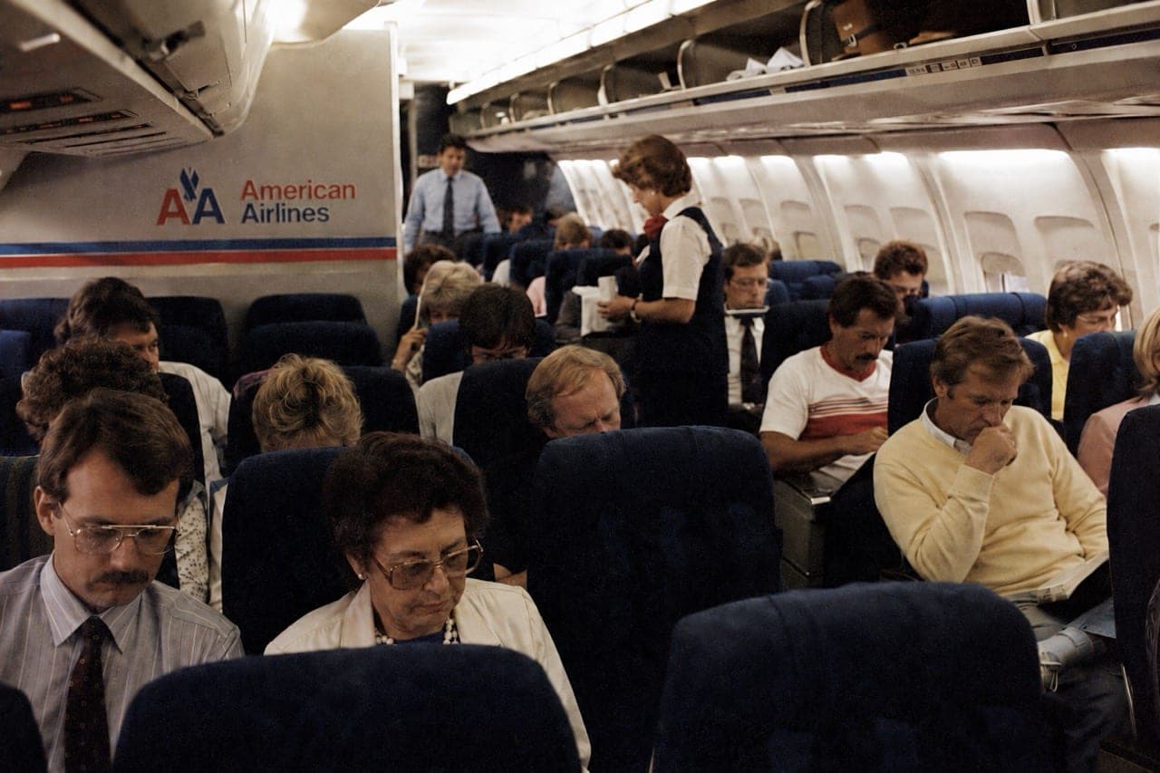 Interior de un avión de American Airlines en los años 80, con pasajeros sentados y una auxiliar de vuelo atendiendo el pasillo.